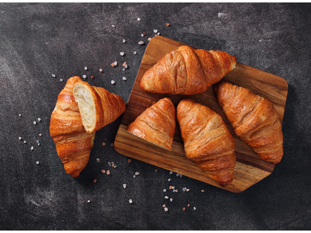 Auksinės spalvos sviestiniai kruasanai, kai kurie sveiki, o vienas perpjautas, ant medinės lentos.