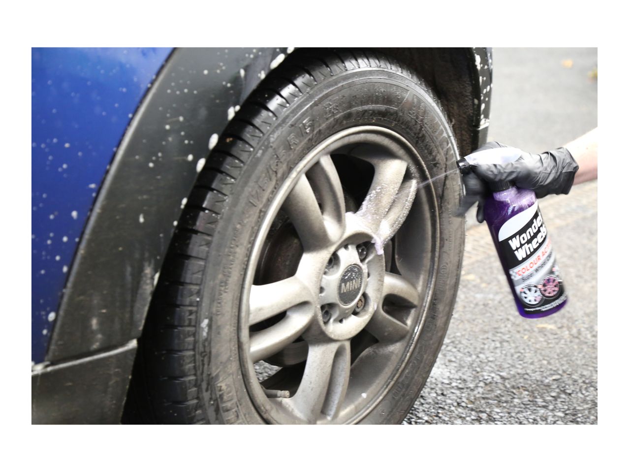 A person in a black glove sprays wheel cleaner onto a dirty car wheel.