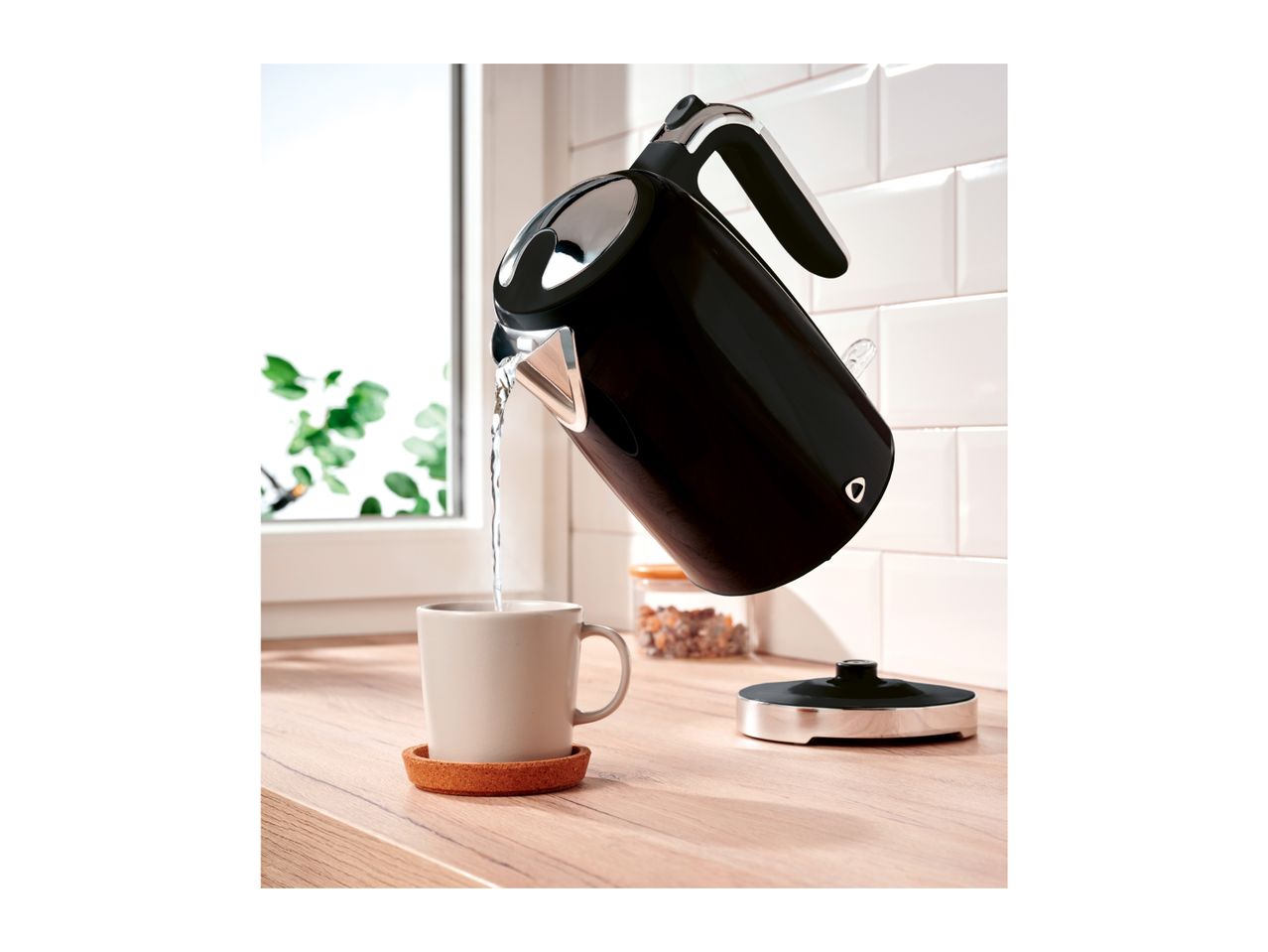 A black electric kettle pouring hot water into a mug on a wooden countertop.