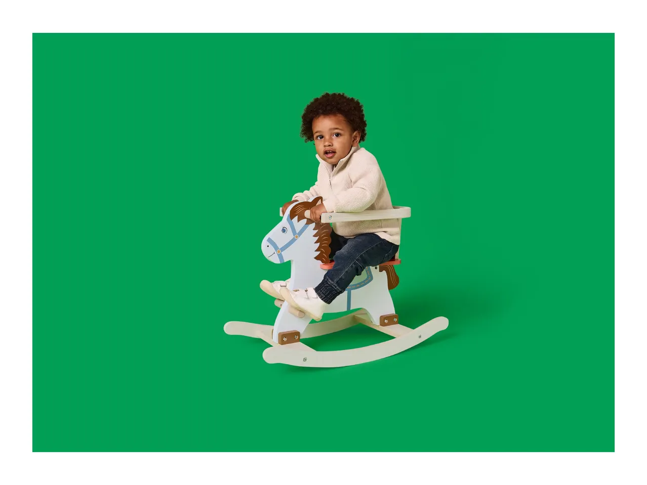 Toddler with curly hair on a white and blue rocking horse against a green background.