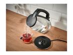 Glass kettle pouring hot water into a cup of tea on a wooden counter.