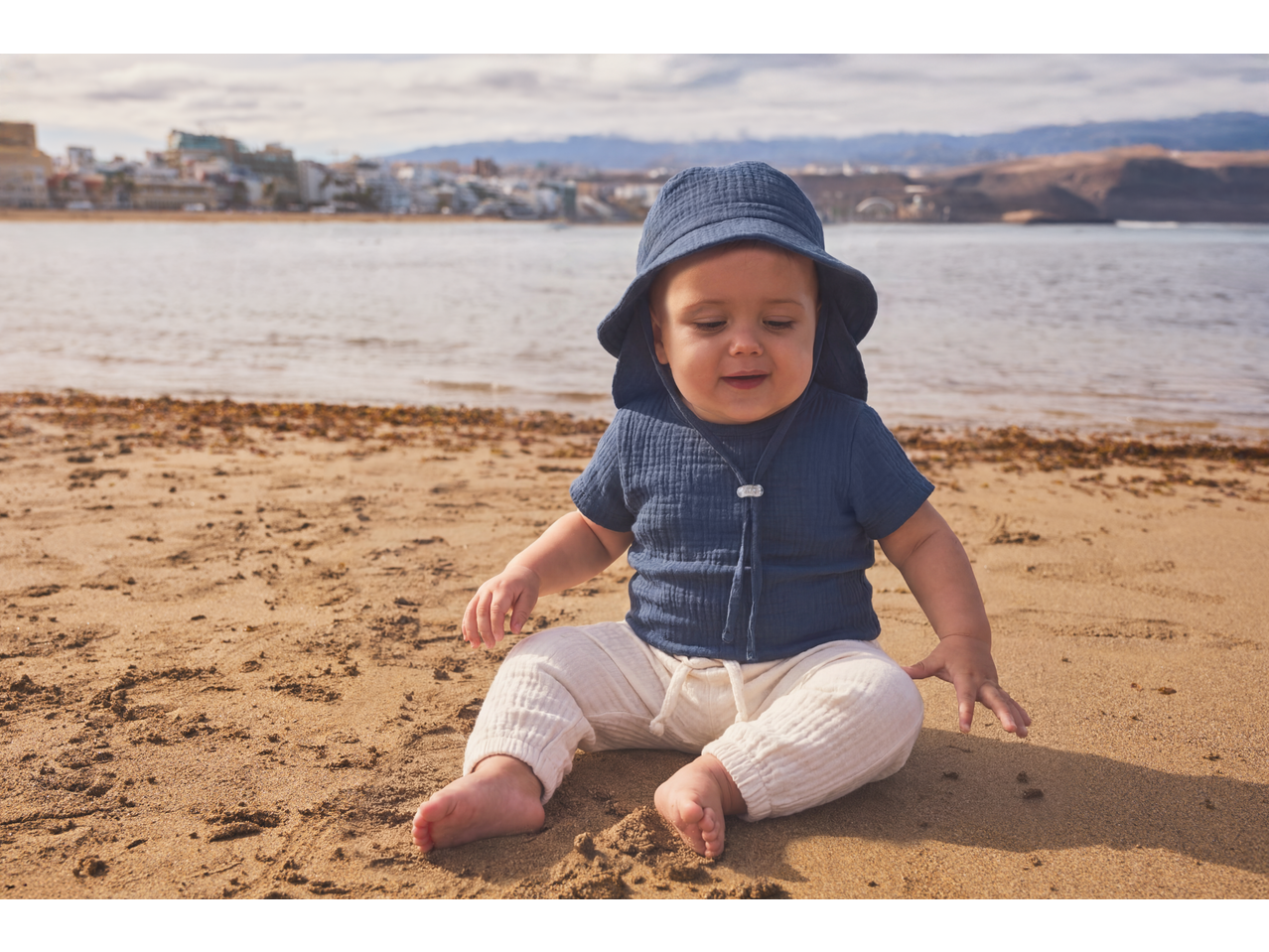 Bebeluș în body de muselină Lupilu și pălărie de soare, așezat pe plajă.