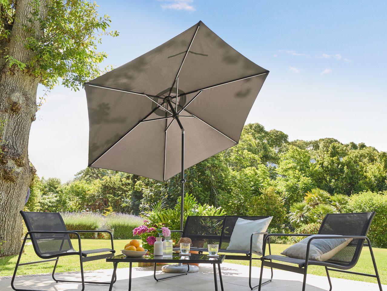 Outdoor patio set with a dark grey umbrella, two chairs, a bench, and a table with fruit and drinks.