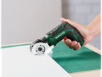 Mão segurando um cortador rotativo sem fio, cortando material branco numa base verde.