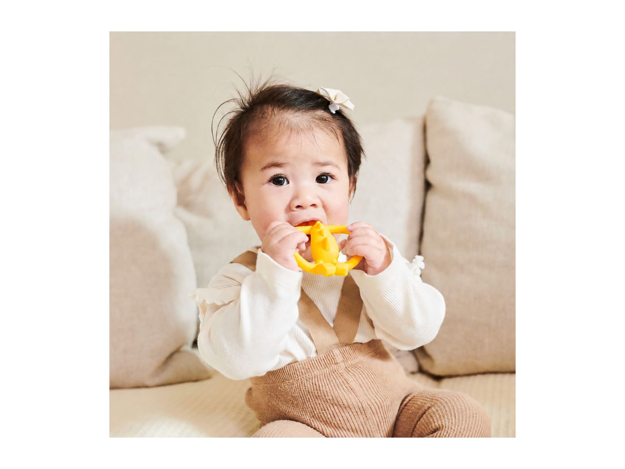 A baby in a white top and brown overalls chews on a yellow teether.