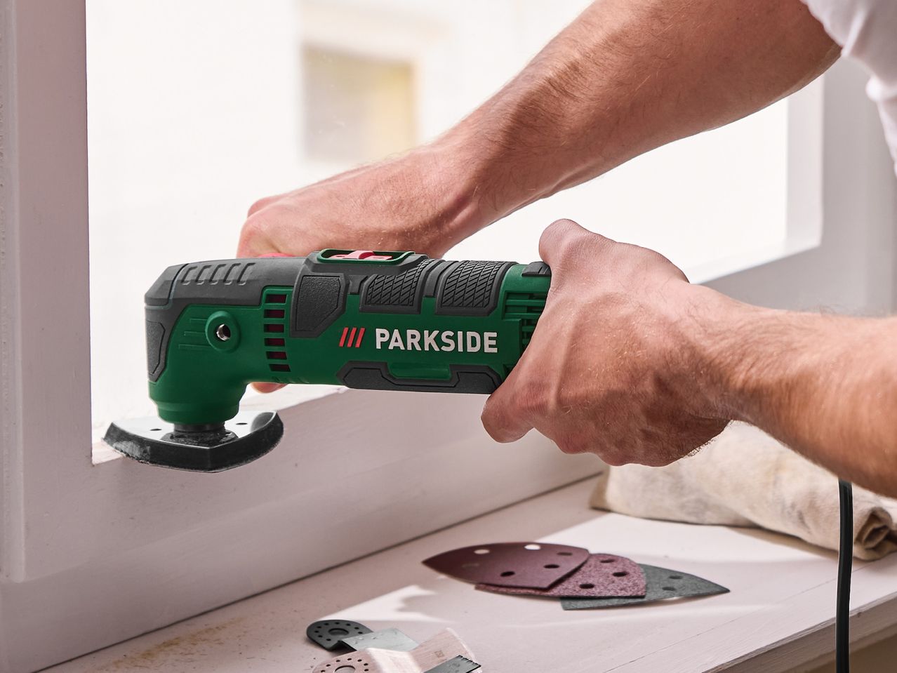 Person sanding a window frame with a green oscillating multi-tool.