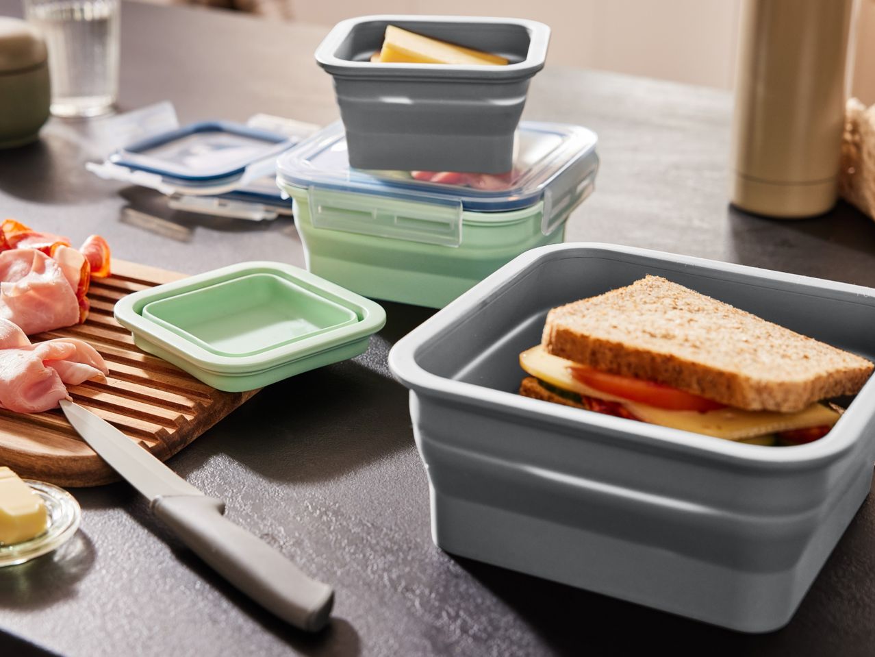 Collapsible food containers with a sandwich, cheese, and deli meats on a counter.