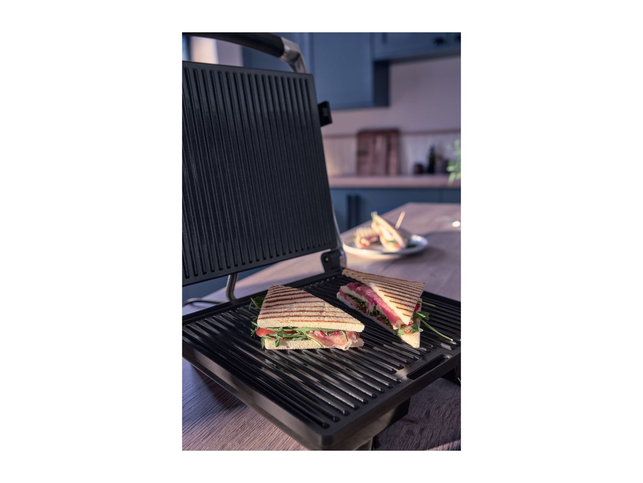 Two grilled sandwiches with prosciutto and arugula on a sandwich maker in a kitchen.