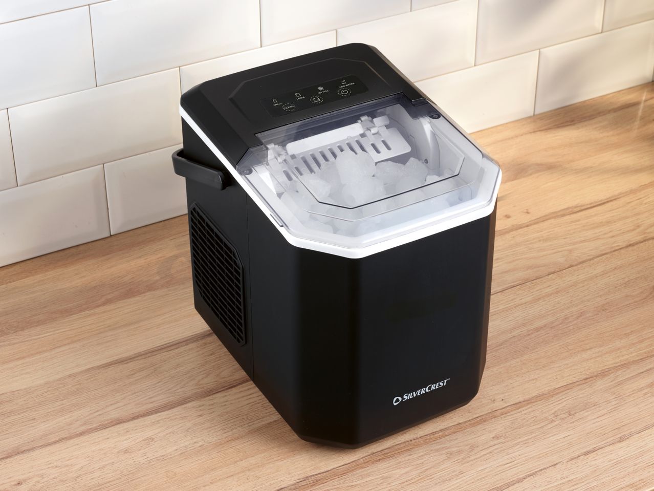 Black ice maker with a clear lid, full of ice, on a wooden countertop next to white tiles.