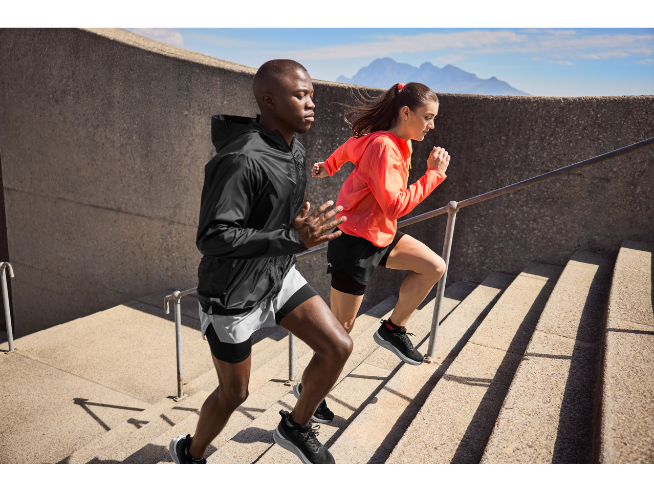 Two athletes in activewear running up outdoor stairs with mountains in the background.