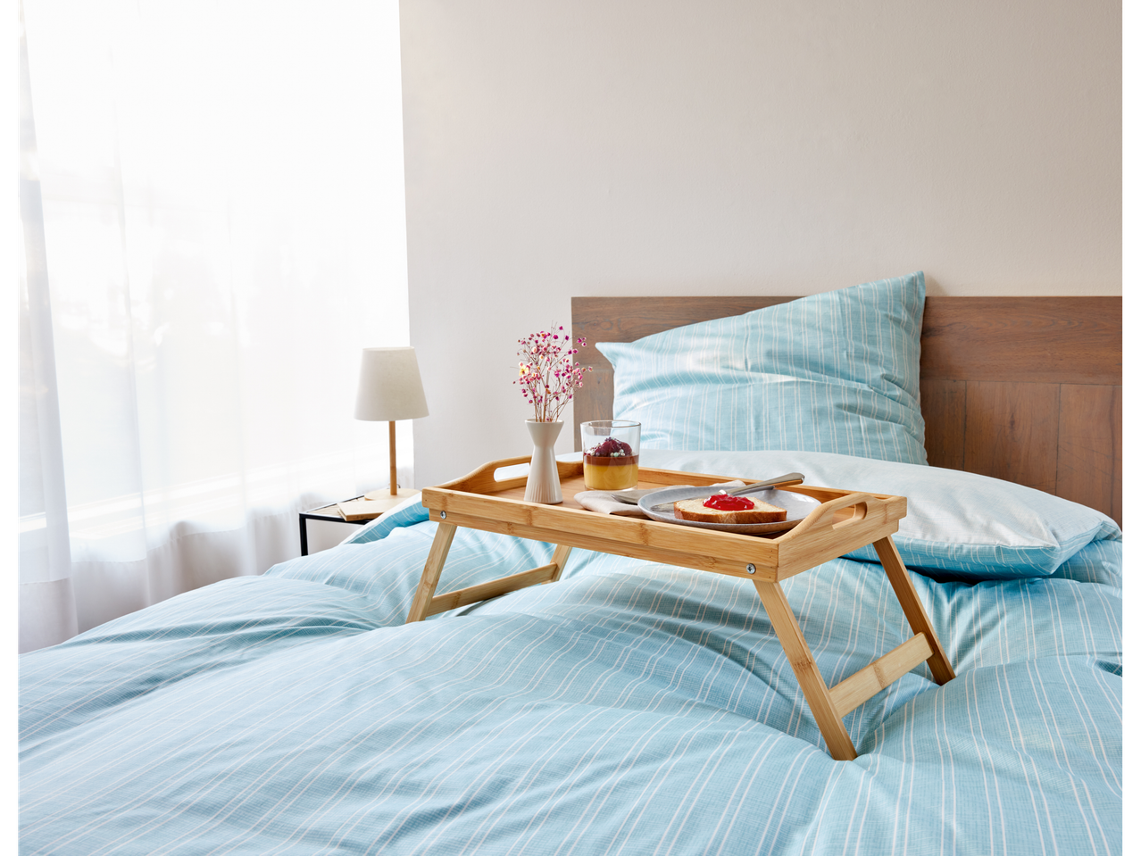 A bamboo breakfast tray sits on a bed with a light blue striped duvet, holding a dessert, toast, and flowers.