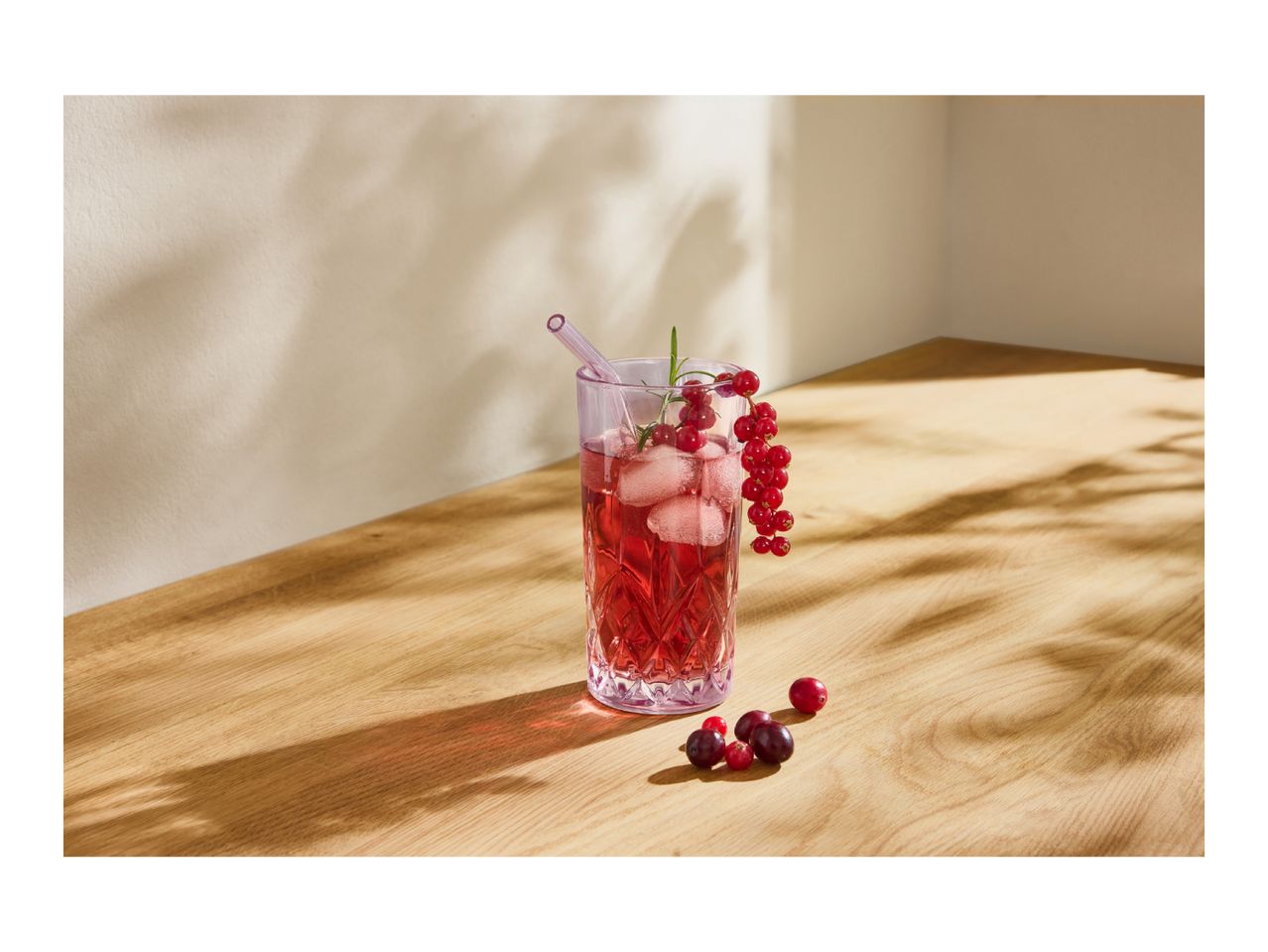 Red berry drink with ice, garnished with redcurrants and rosemary, on a wooden surface.
