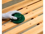 Gloved hand sanding wooden slats with a green manual sander.