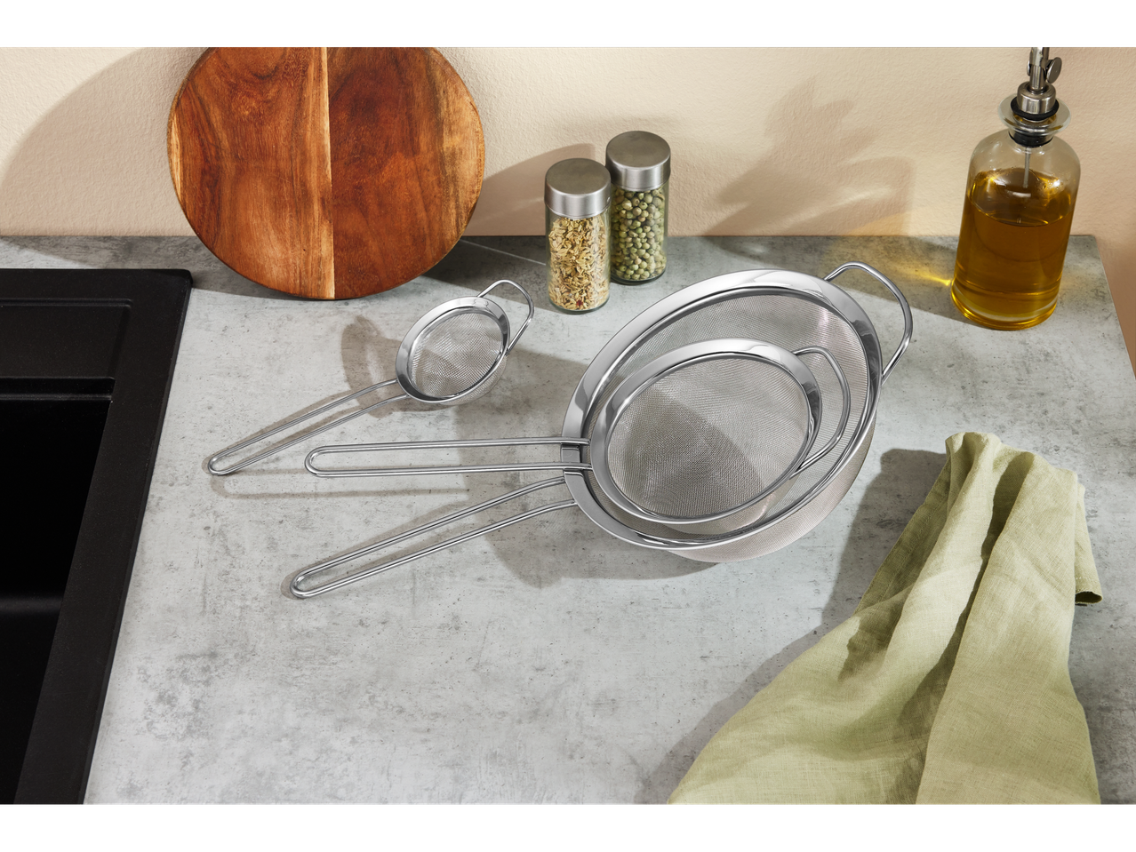 Kitchen counter with stainless steel strainers, cutting board, spice jars, and oil bottle.
