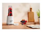 A white personal blender filled with berries next to a bowl of fresh fruit on a wooden counter.