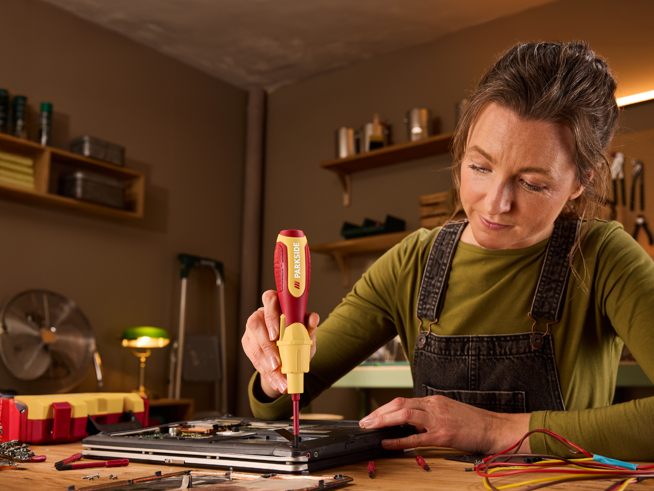 Жена поправя лаптоп с отвертка, на маса с инструменти и кабели.