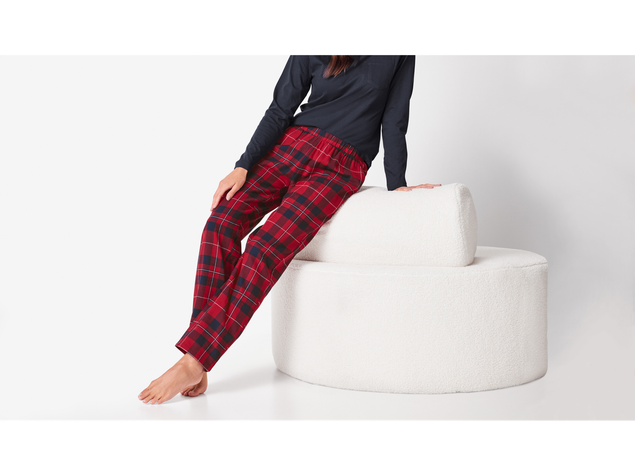 Esmara: Femeie în pantaloni de pijama în carouri roșii și negre, așezată pe un puf alb.