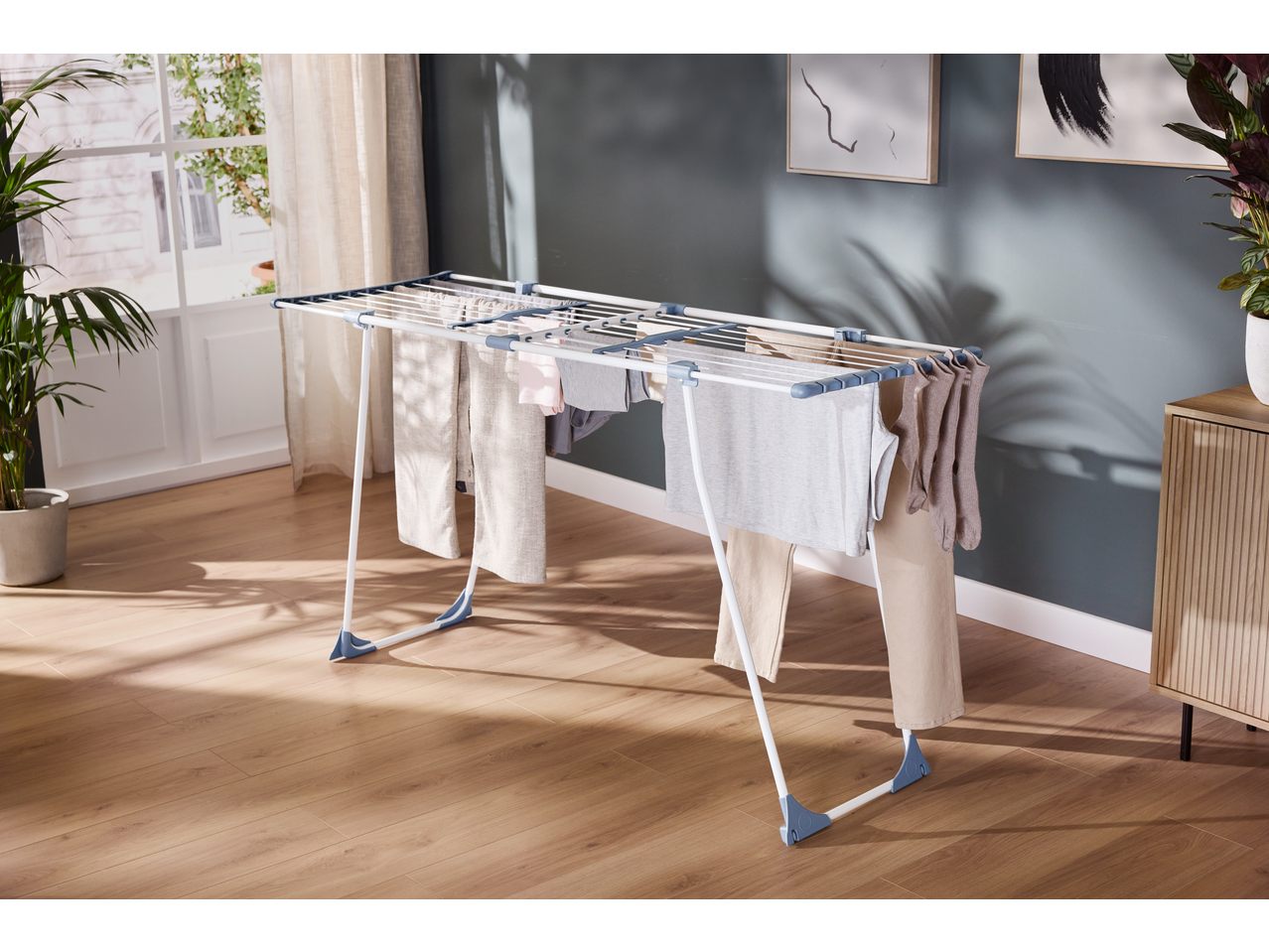 Clothes drying rack with various garments in a bright room with wooden floor and plants.