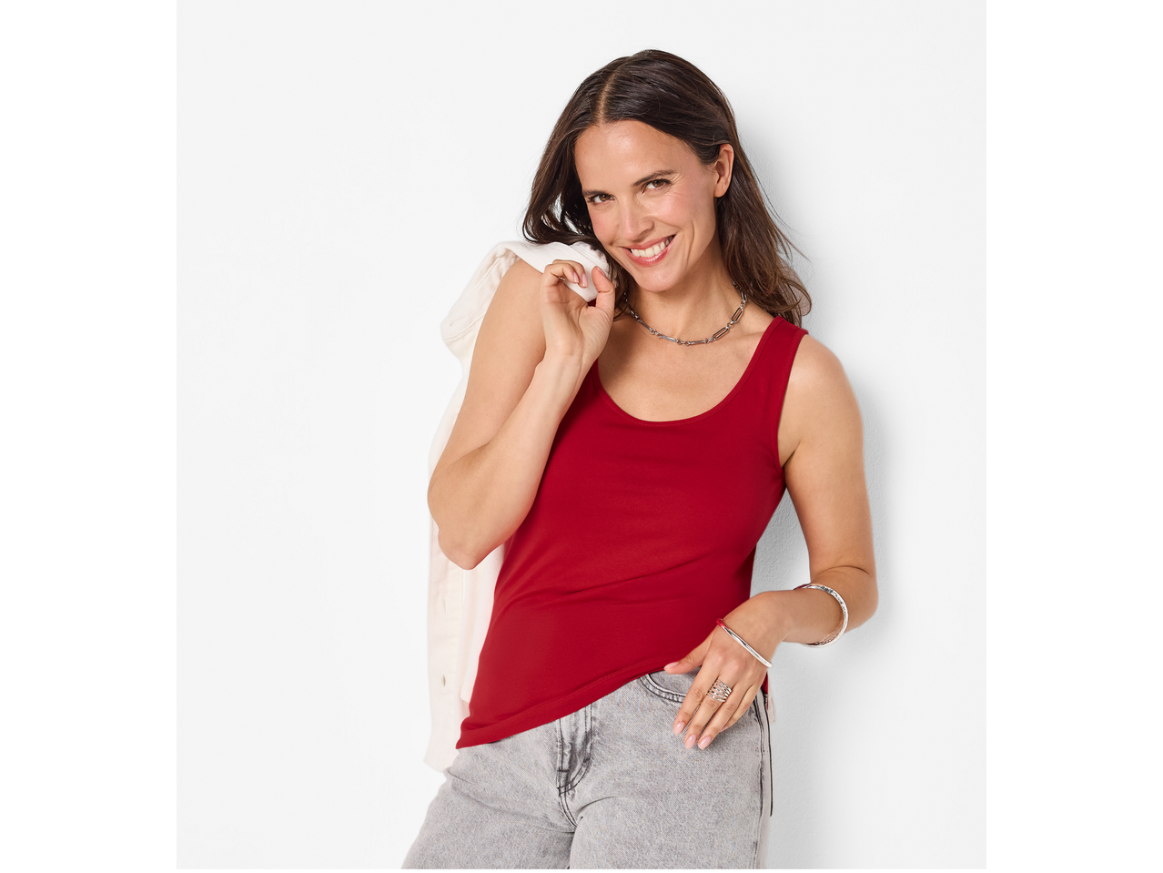 Nasmejana žena u crvenom ESMARA topu i sivim farmerkama, sa belom jaknom na ramenu.