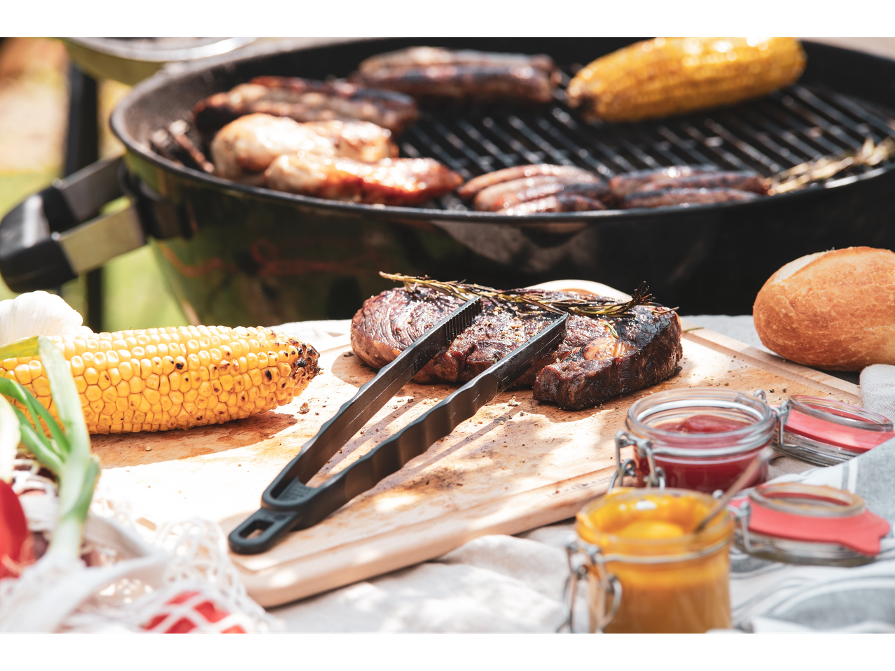 Grilled meat, sausages, and corn on a barbecue with condiments and bread.