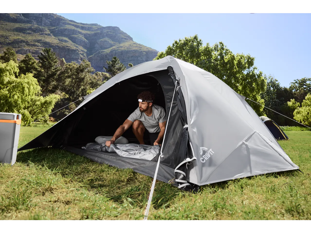 Man in CRIVIT 4 Person Blackout Tent, setting up sleeping bag, mountains in background.