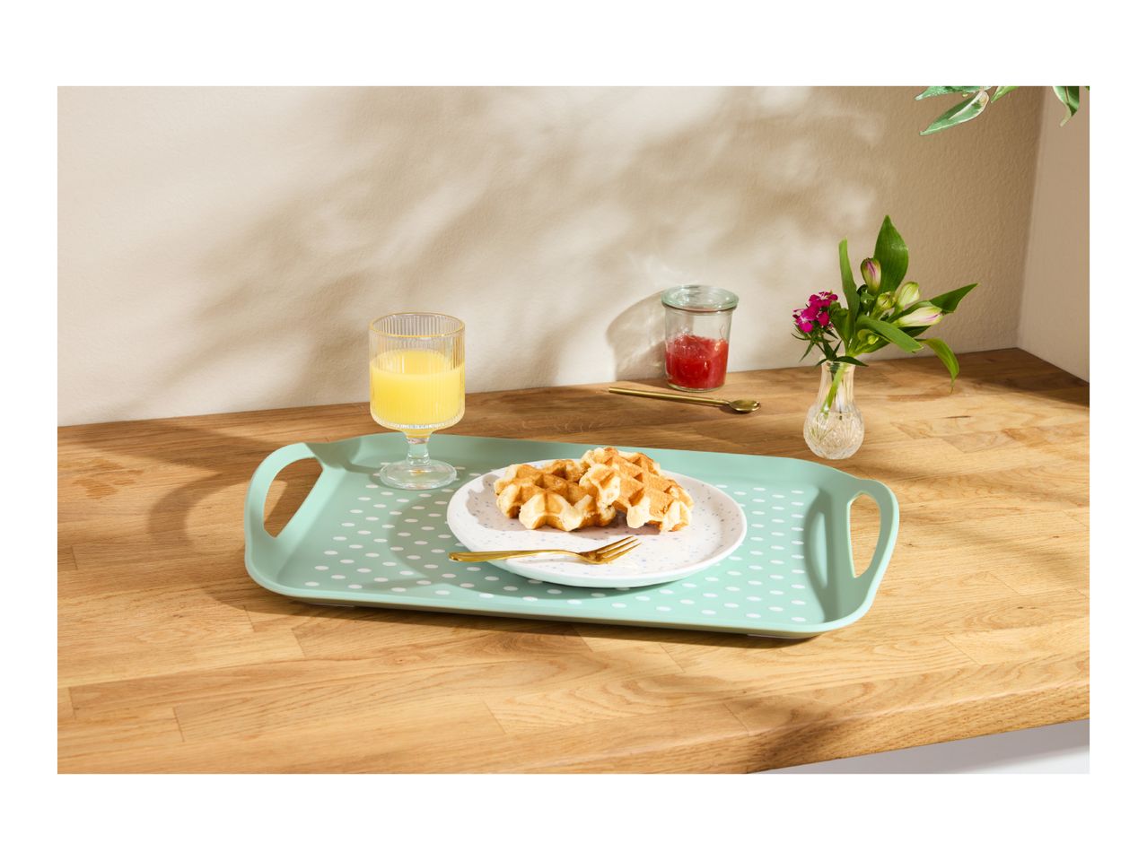 Breakfast tray with waffles, orange juice, jam, and flowers on a wooden counter.