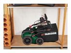 Green lawnmower with a black grass collection bag on a wooden shelf in a storage area.