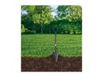 Shovel digging into soil with grass and trees in the background.