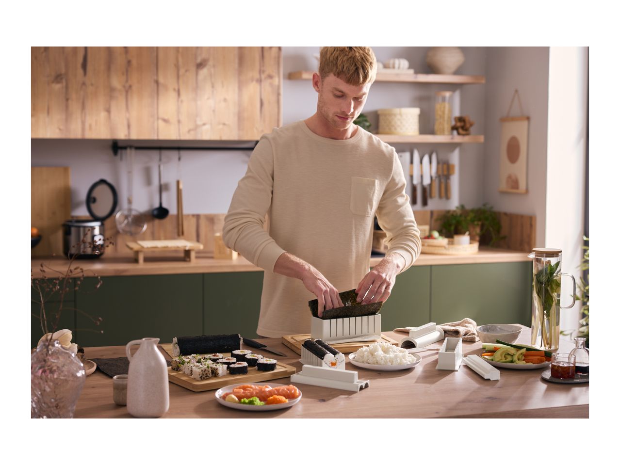 Man making sushi with a sushi maker, rice, salmon, and vegetables on a wooden table.