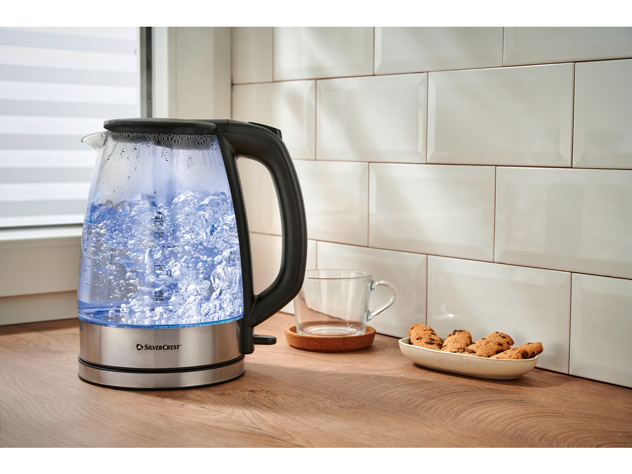 SILVERCREST® Colour Changing Glass Kettle boiling water on a kitchen counter with cookies.
