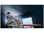 Homme utilisant une scie circulaire pour couper une planche de bois, avec de la poussière en suspension.