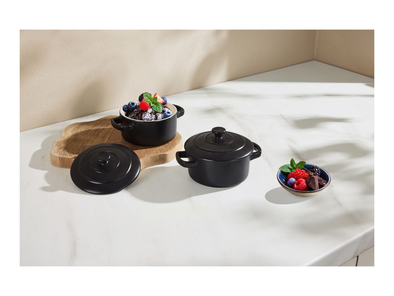 Black ceramic mini cocottes with lids, one filled with chocolate cake and berries, on a wooden board.