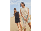 Two women in casual summer tops and shorts, one holding a net bag with fruit, on a sandy beach.