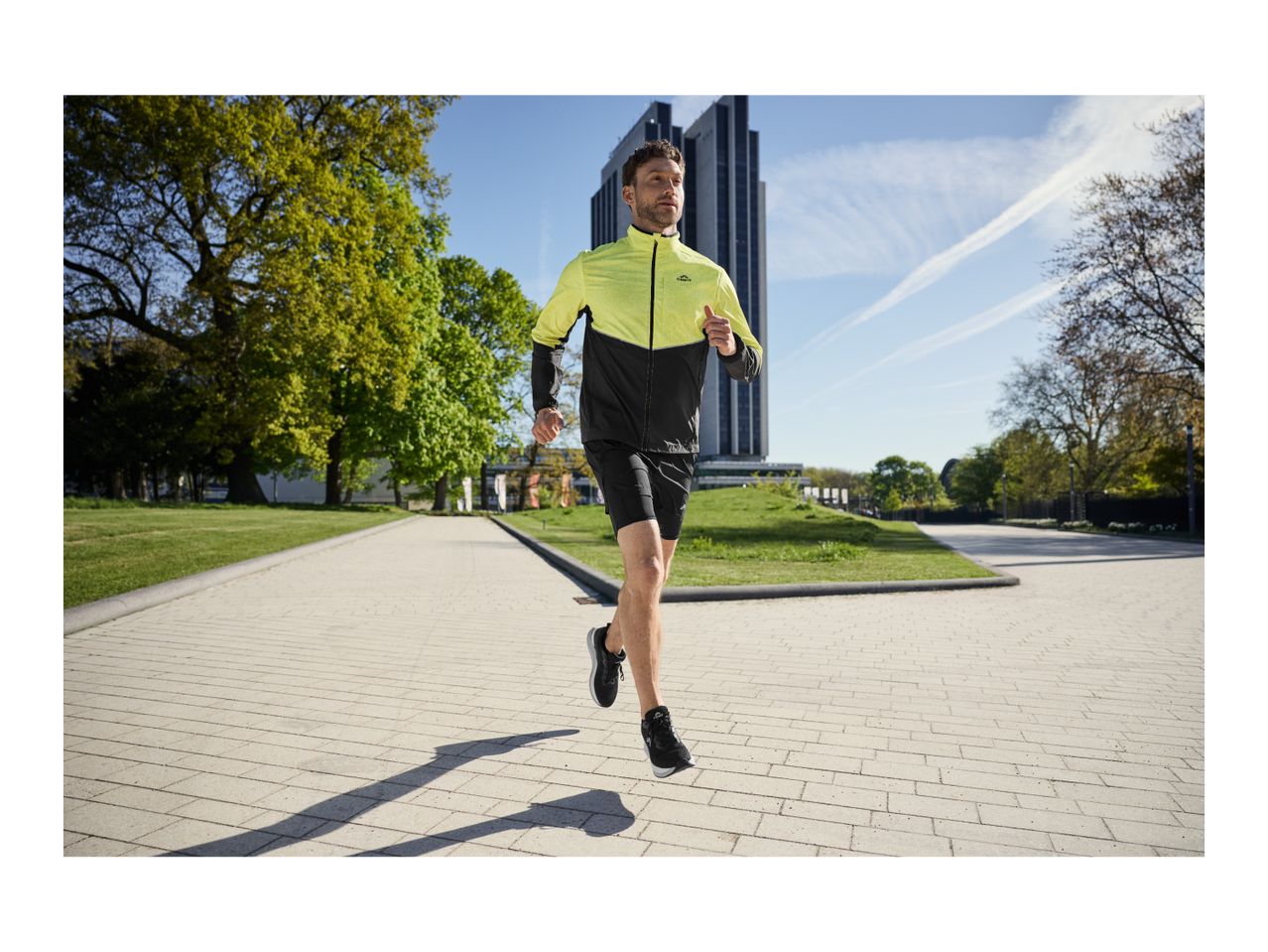 Man running in a black and neon yellow sports jacket and black shorts on a paved path.