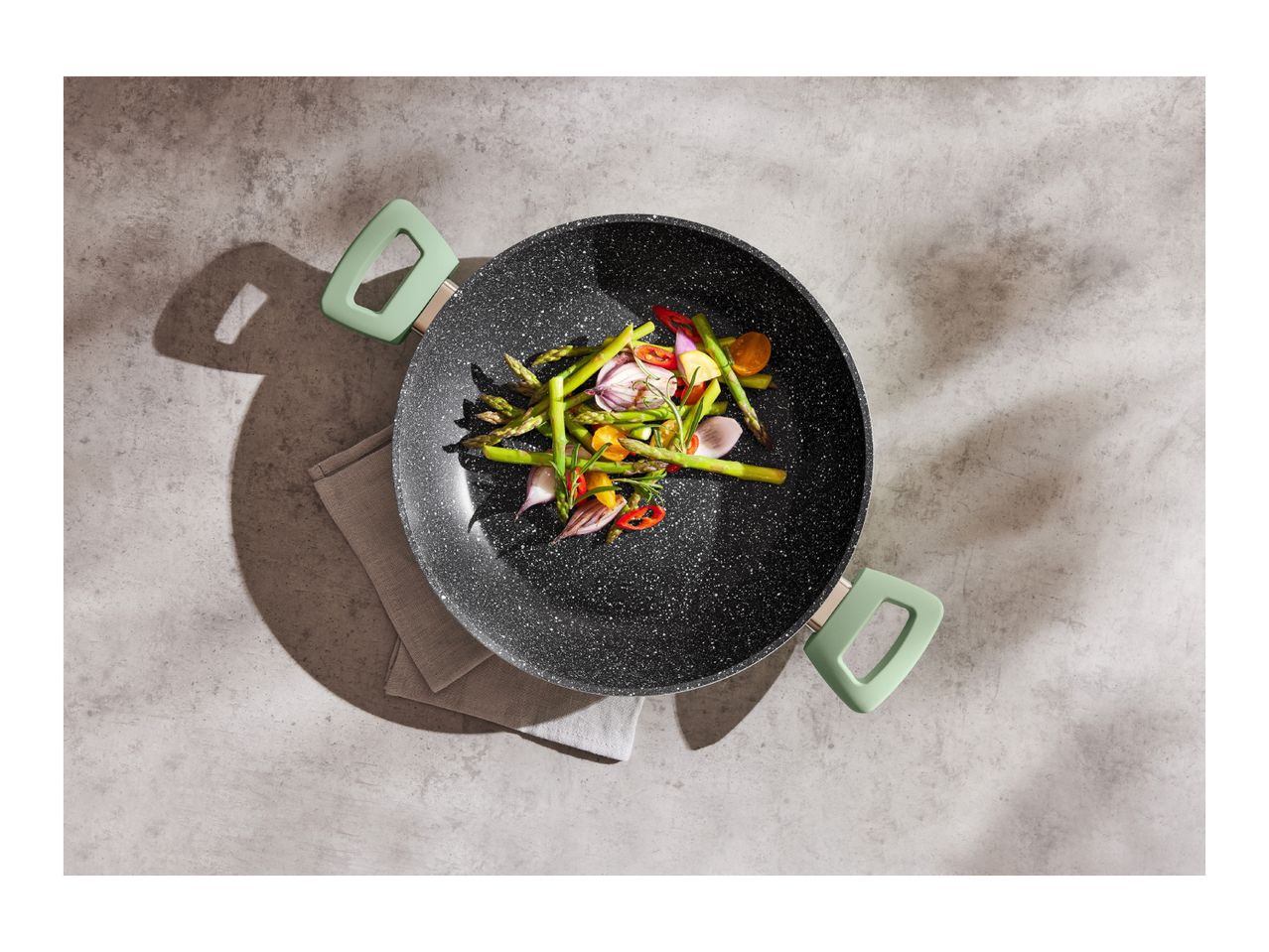 Black wok with green handles filled with stir-fried vegetables on a grey surface.