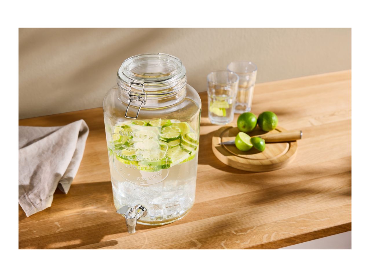 Glass drink dispenser with lime slices, two glasses, and limes on a wooden board.
