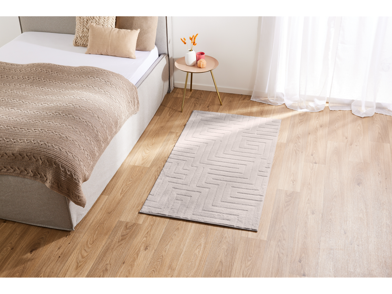 A bedroom with a bed, a knitted blanket, a side table with decor, and a patterned rug on a wooden floor.