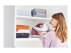 Woman storing a steam generator iron in a white cabinet with folded clothes and a toiletry bag.