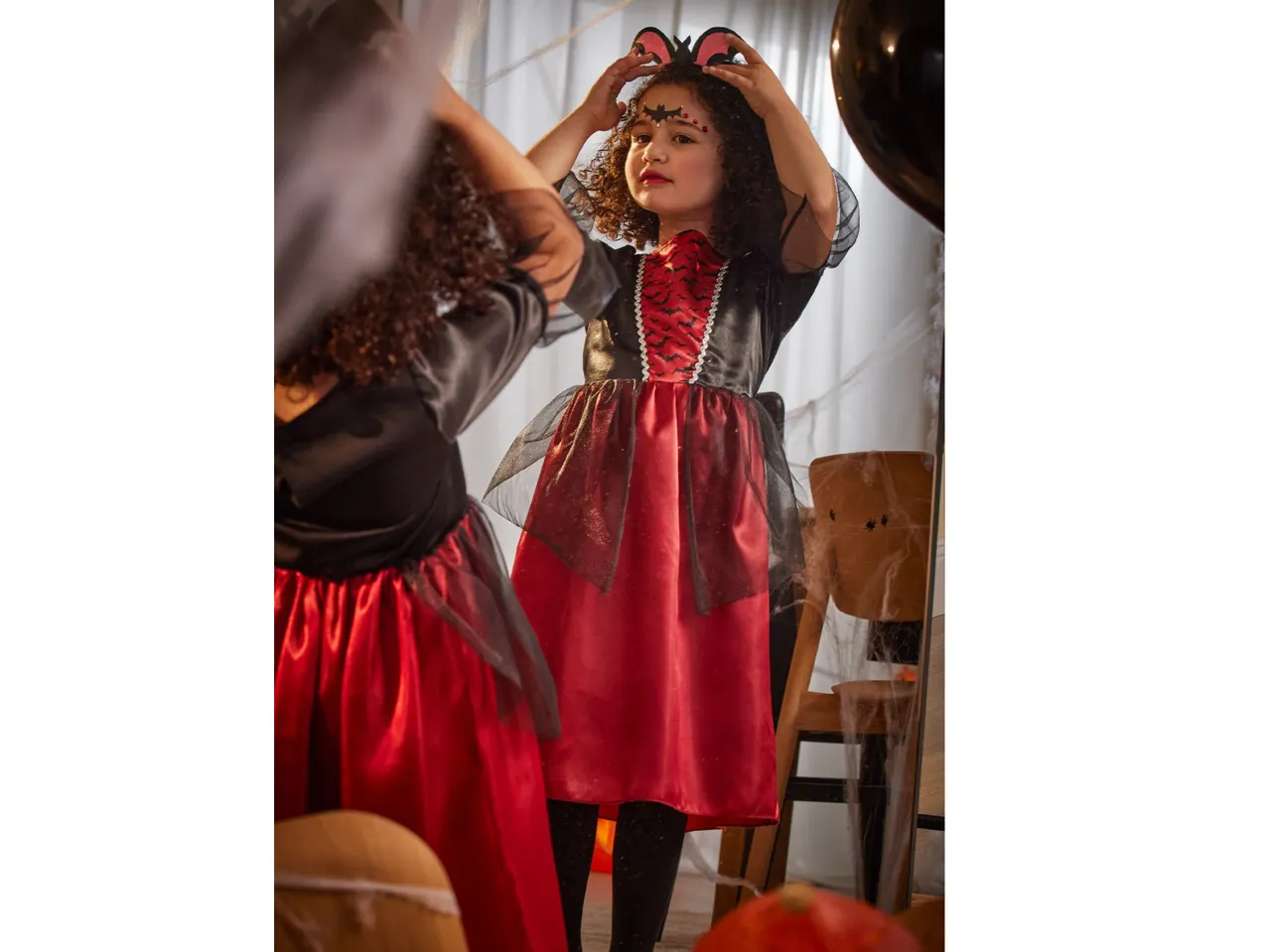 A girl in a Halloween bat costume with face paint looks in a mirror.