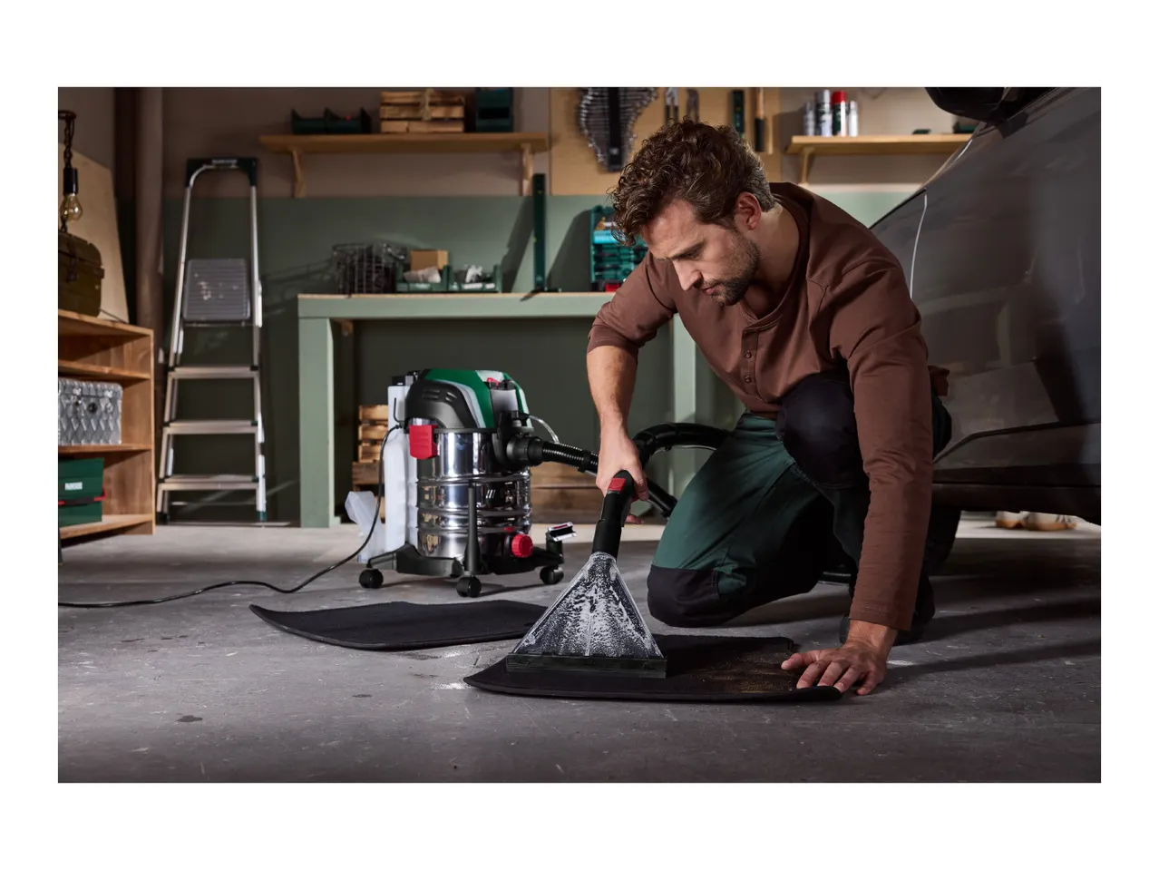 A man cleaning a car mat with a wet and dry vacuum cleaner in a garage.