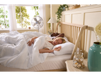A couple sleeps in a bed with white bedding and a fan.