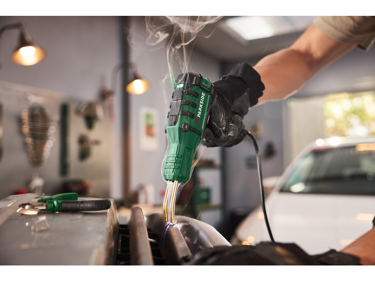 Persoană care sudează cu un pistol de sudură plastic verde PARKSIDE, fum ridicându-se, într-un garaj.