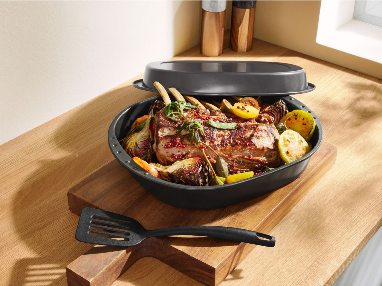 Печено месо с гарнитура от зеленчуци в овален съд за печене с капак и шпатула.