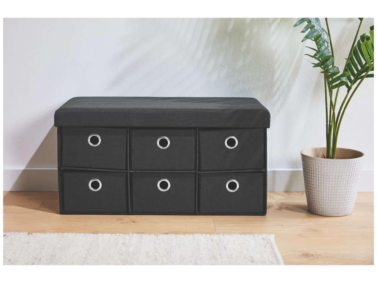 Un banc de rangement noir avec six tiroirs et un coussin rembourré, à côté d'une plante en pot.