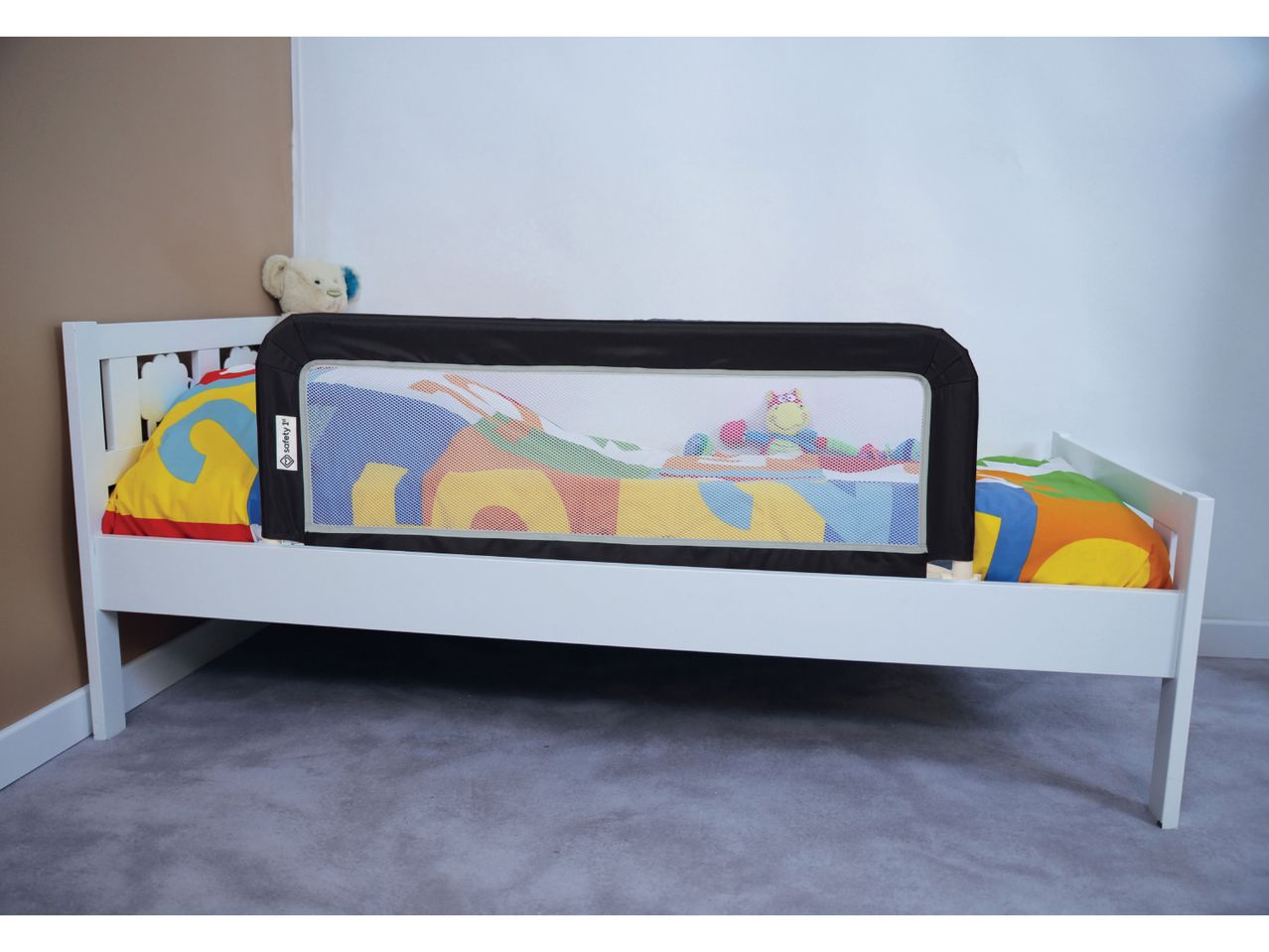 White toddler bed with a black and mesh bed rail, colorful bedding, and a teddy bear.