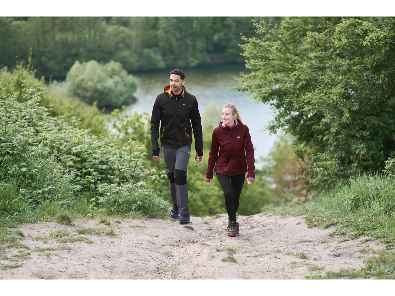 Two people in CRIVIT sports tops walking on a dirt path near a lake.
