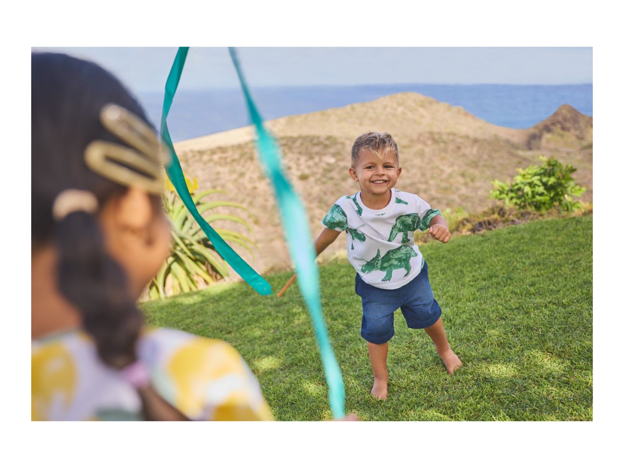 A boy in a dinosaur t-shirt and denim shorts plays with a ribbon outdoors.