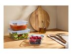 Glass food containers with soup, salad, and berries on a wooden counter with a cutting board.