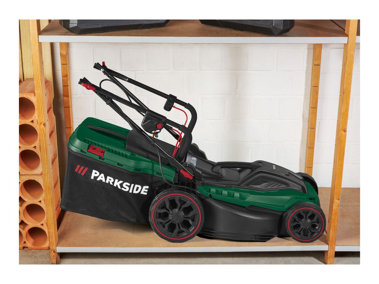 A green and black lawnmower with a grass collection bag on a wooden shelf.