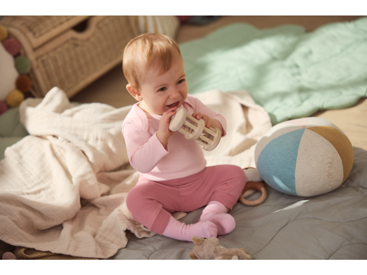 Happy baby in pink clothes playing with a wooden rattle on a soft blanket.